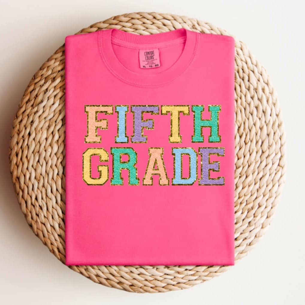 Pink t-shirt with 'FIFTH GRADE' in colorful letters on a woven mat.