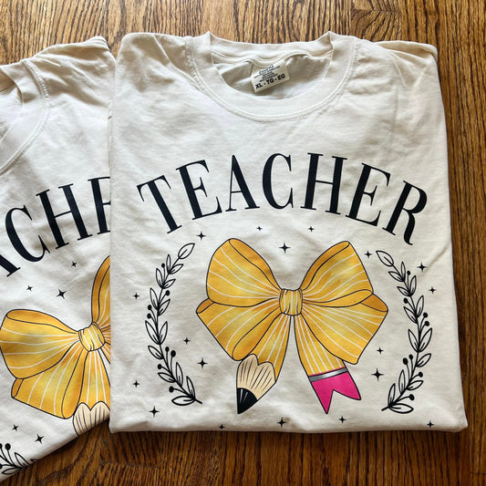 Two folded t-shirts with 'TEACHER' text and a yellow bow design on a wooden surface.