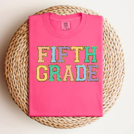 Pink t-shirt with 'FIFTH GRADE' in colorful letters on a woven mat.